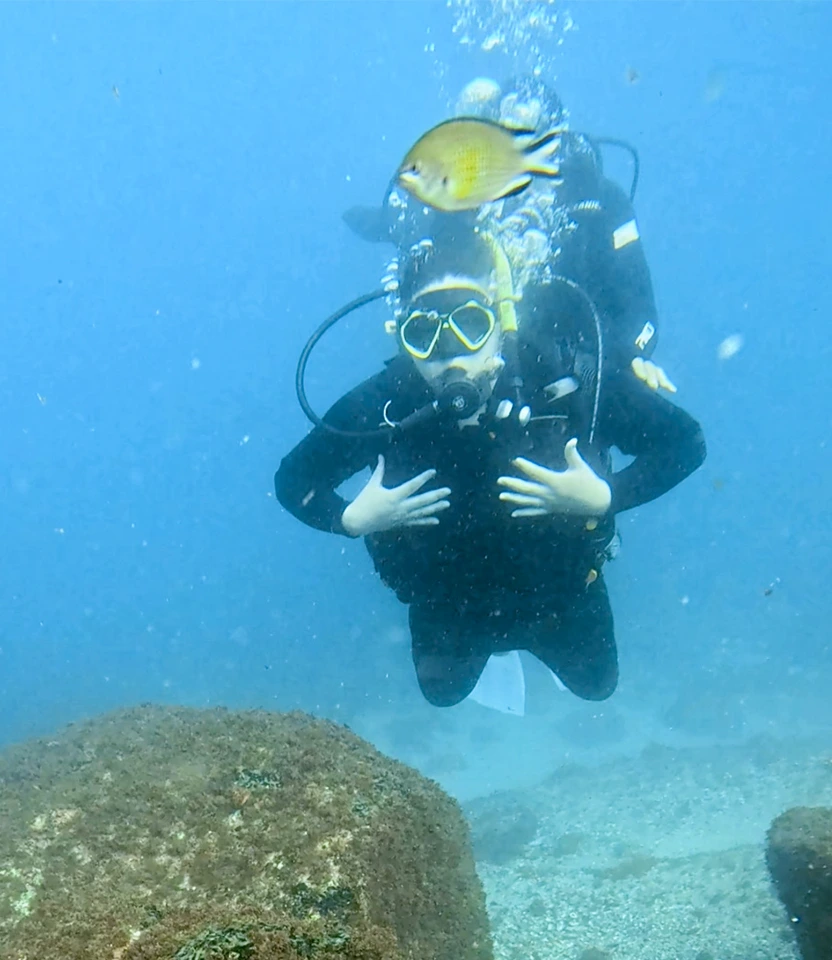 Mergulhador fazendo sinal de ok no fundo do mar com peixe amarelo nadando à frente.