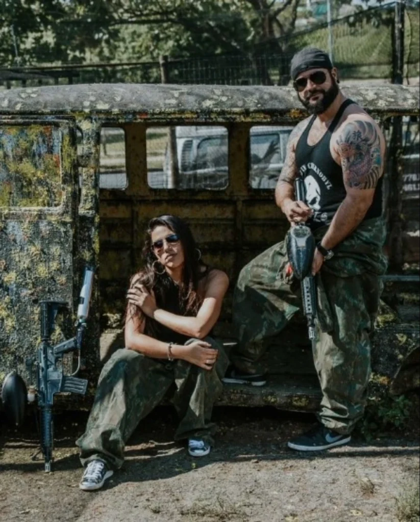 Ensaio fotográfico de casal de mãos dadas e algemados segurando marcadores de paintball em cenário temático em Florianópolis.