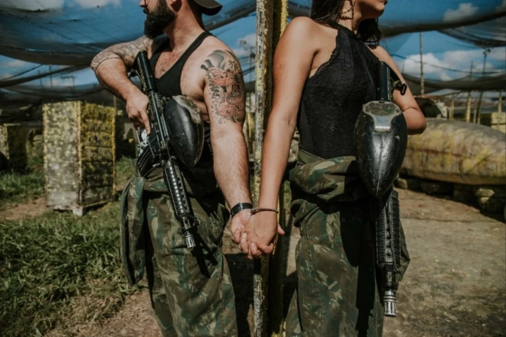 Casal de óculos escuros e trajes camuflados em ensaio fotográfico conceitual com marcadores de paintball e kombi abandonada em Florianópolis.