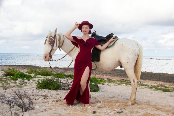 Mulher com vestido elegante vinho posando ao lado de cavalo branco na praia em Florianópolis.