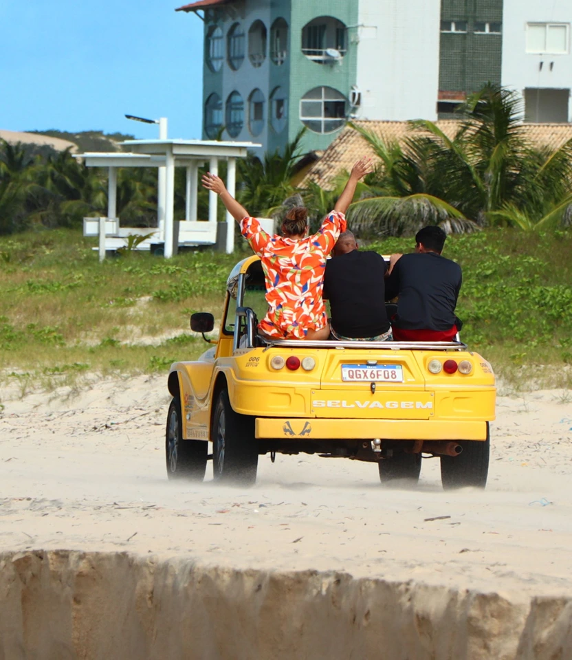 Buggy amarelo "Selvagem" transitando pelas areias da praia em Florianópolis com turista com braços abertos.
